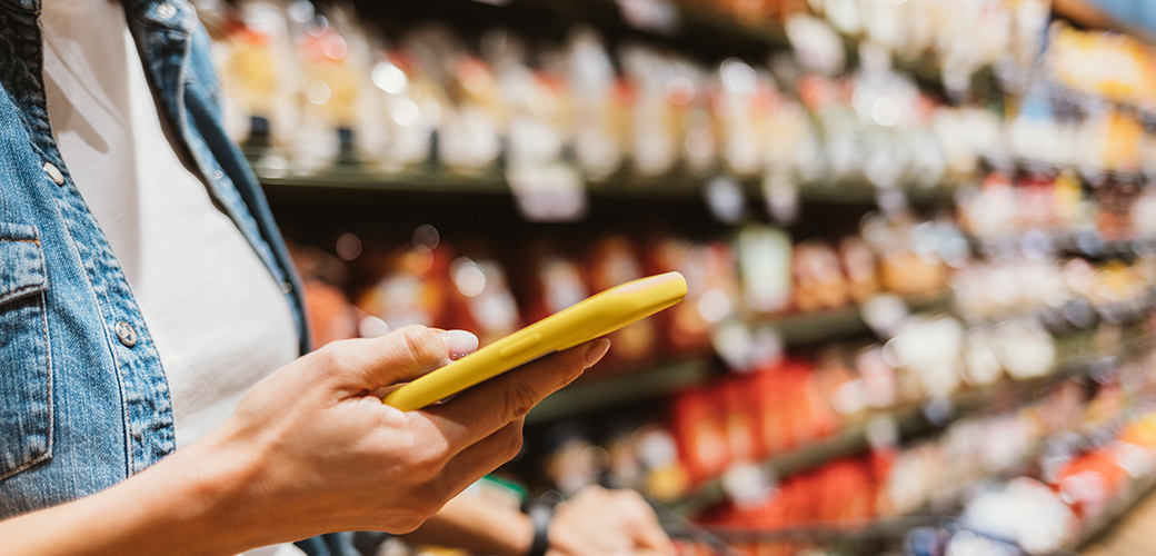 Close up on a woman's hand holding a phone in yellow case while standing a grocery store aisle